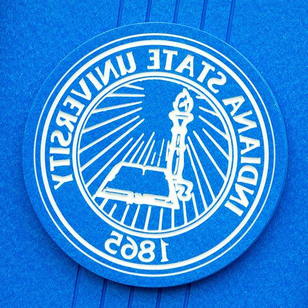 The Indiana State University seal. It is white on a blue background and features a torch, an open book, and the year 1865.