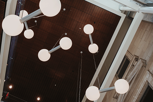 Art installation featuring white and gray art pieces that look like barbells, suspended from the ceiling in the Health and Human Services Building. The five sets of round white end pieces and the gray connectors stand out against the dark ceiling above.