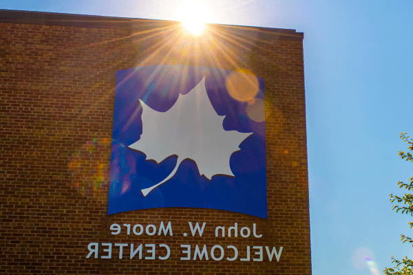 A large white Sycamore leaf logo on a blue background is visible above the name of the John W. Moore Welcome Center. The sun is peeking just above the red brick building, casting rays of sunshine over the top of the logo.