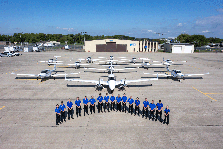 A group of people in matching blue polo shirts and black trousers stands in a curved formation on a large concrete tarmac. Behind them, a fleet of small white aircraft is neatly arranged. A beige hangar with brown doors and a blue sign stands in the background under a blue sky, surrounded by trees, buildings, and parked vehicles.  