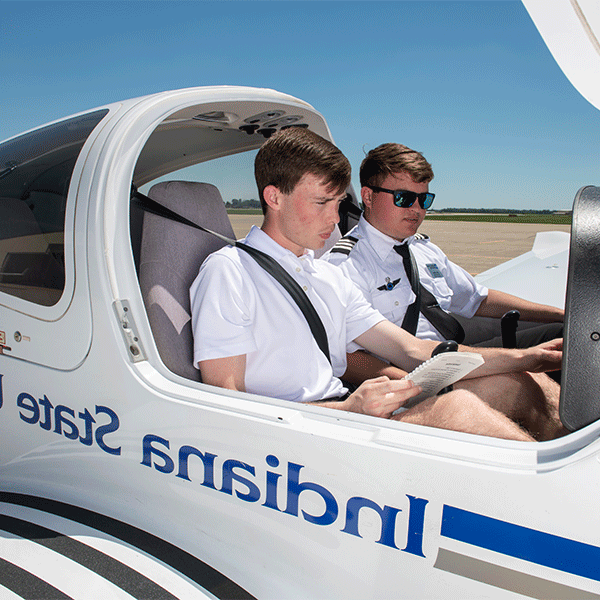 Two young white men with dark brown hair are seated inside an aircraft with an open canopy. One, wearing a white polo shirt, holds a checklist, while the other, dressed in a pilot's uniform and sunglasses, observes. Both are wearing seatbelts. The side of the aircraft displays “Indiana State” in blue. The background features a clear blue sky and a tarmac.