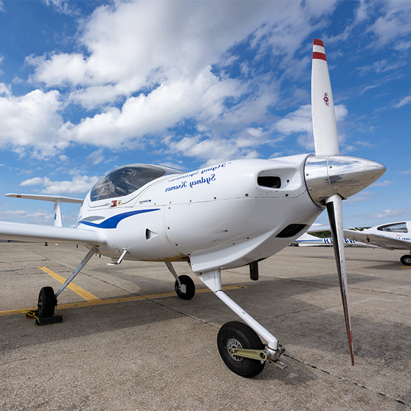 A white propeller aircraft with blue letters reading “Flying Sycamores Sydney Kremer” on its body sits on a tarmac under a bright blue sky with scattered clouds. Another white aircraft is partially  visible on the left  side.