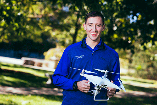 A smiling white man with short dark hair stands outdoors with greenery visible behind him. He wears a blue zip-up jacket that says “Indiana State University” and holds in both hands a white drone with three propellers on it.  