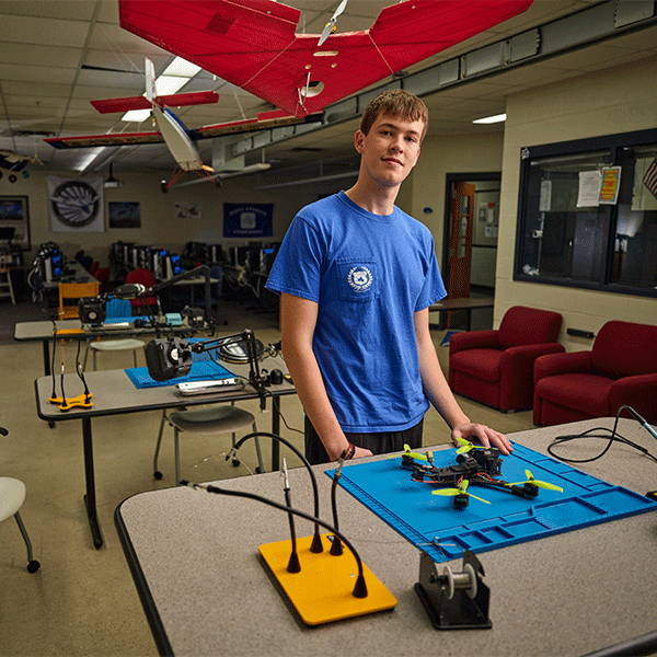 A young white man with short light brown hair in a blue t-shirt stands in a classroom, resting his hand on a small drone on a blue mat. He is smiling slightly and exudes confidence. The room contains various tools and workstations. Several model aircraft with red wings hang from the ceiling. The background shows computers, workstations, and red lounge chairs.