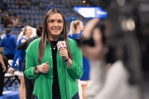 A white female student with long brown hair is at a basketball game inside the Hulman Center. She wears a green sweater vest with a black undershirt. She holds a microphone with a red “E” on a white square on the microphone. She is looking toward a cameraperson holding a black camera. Cheerleaders in blue uniforms are visible behind her. The arena’s bleachers are also visible in the background. 