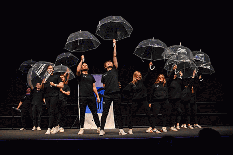 A group of male and female students dressed in black stand illuminated on a dark stage as they raise clear plastic umbrellas above their heads. A blue-and-white banner is visible behind them. One man on the right side holds his umbrella at a left angle to the side and not above his head like the others.