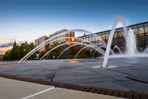 Low angle of a fountain with streams of water in the foreground and the Welcome Center and Sycamore Towers and blue sky in the background. 