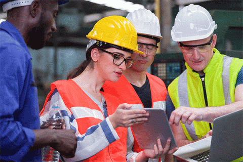 Diverse group of workers wearing hard hats and safety vests look at data on a tablet in a factory setting. 