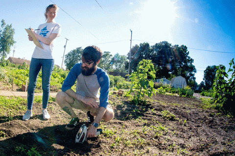 A MALE STUDENT KNEELS ON THE GROUND AND CHECKS THE SOIL WITH AN INSTRUMENT WHILE A FEMALE STUDENT STANDING NEARBY TAKES NOTES IN A GARDEN SETTING. 