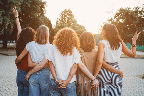 A group of five women are seen standing from the back. They are holding hands and have their arms around each other as they look toward a sunset framed by green trees in the background. The women wear an assortment of jeans, t-shirts, and other summer clothing, and their hair has various styles and colors. 