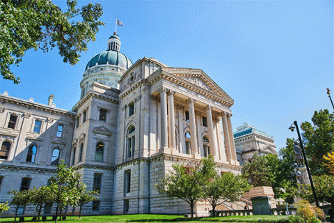 Outside image of Indiana State University’s Normal Hall, a stately white stone building, three stories high, with tall columns, wide steps, and a dome at the top. Blue sky is visible above, and the building is surrounded by green grass and several green trees.